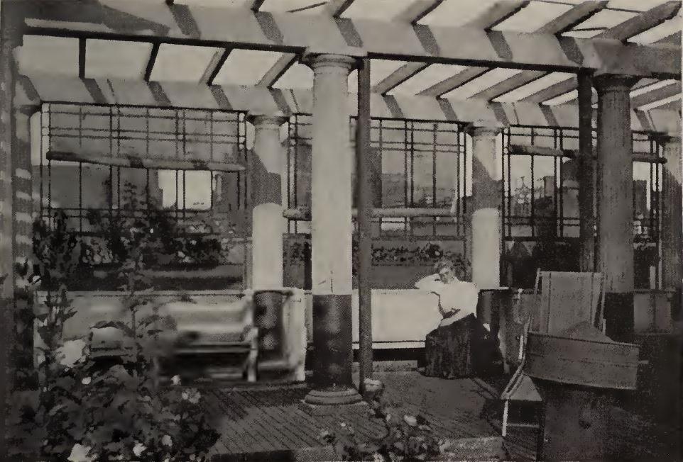 在钱伯斯街44号屋顶花园拍摄的黑白照片. 照片中，一位身穿白衬衫和深色长裙的女士坐在长凳上. 有4个完整的柱子支撑水平梁(一个凉棚)。. 还可以看到几株植物. 