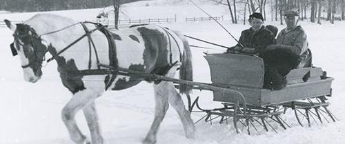 Black and white photograph of a horse pulling a sleigh through snow. 一个男人和一个女人坐在雪橇上.